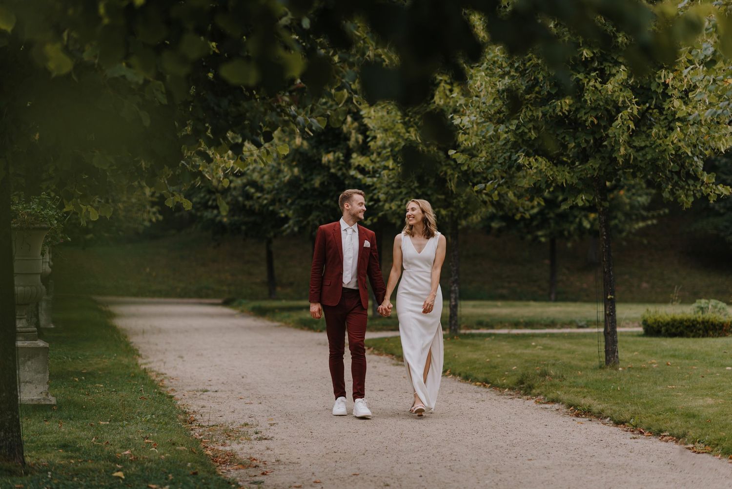 Wedding couple walks along a tree-lined path in maroon and white attire.