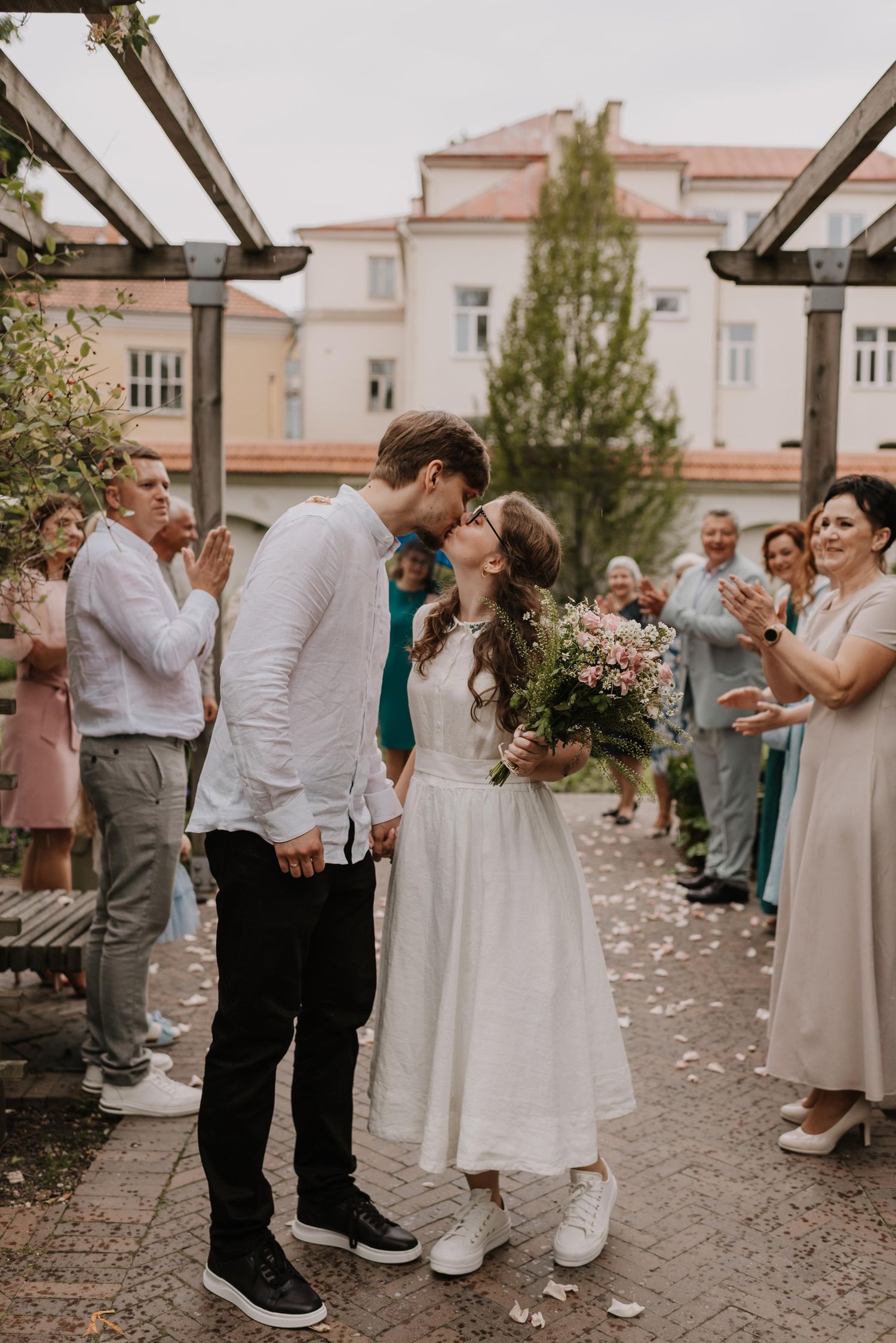 A couple embraces during an outdoor wedding ceremony surrounded by rose petals and guests in light-colored attire.