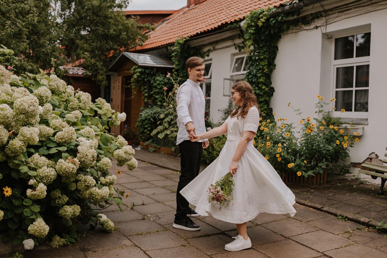 Couple poses outdoors near a white building with climbing roses and hydrangea bushes.