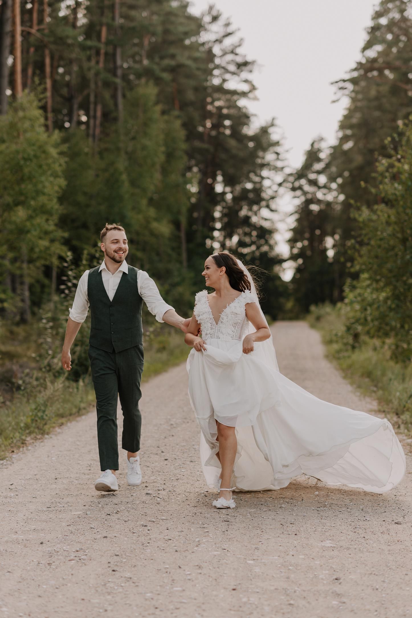 A couple walks along a scenic dirt path through tall pine trees while the bride's dress trails behind.