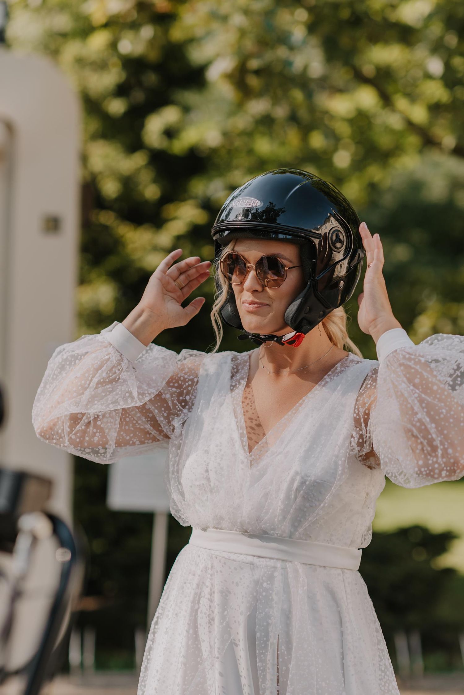 Person in white lace dress and helmet poses playfully outdoors.