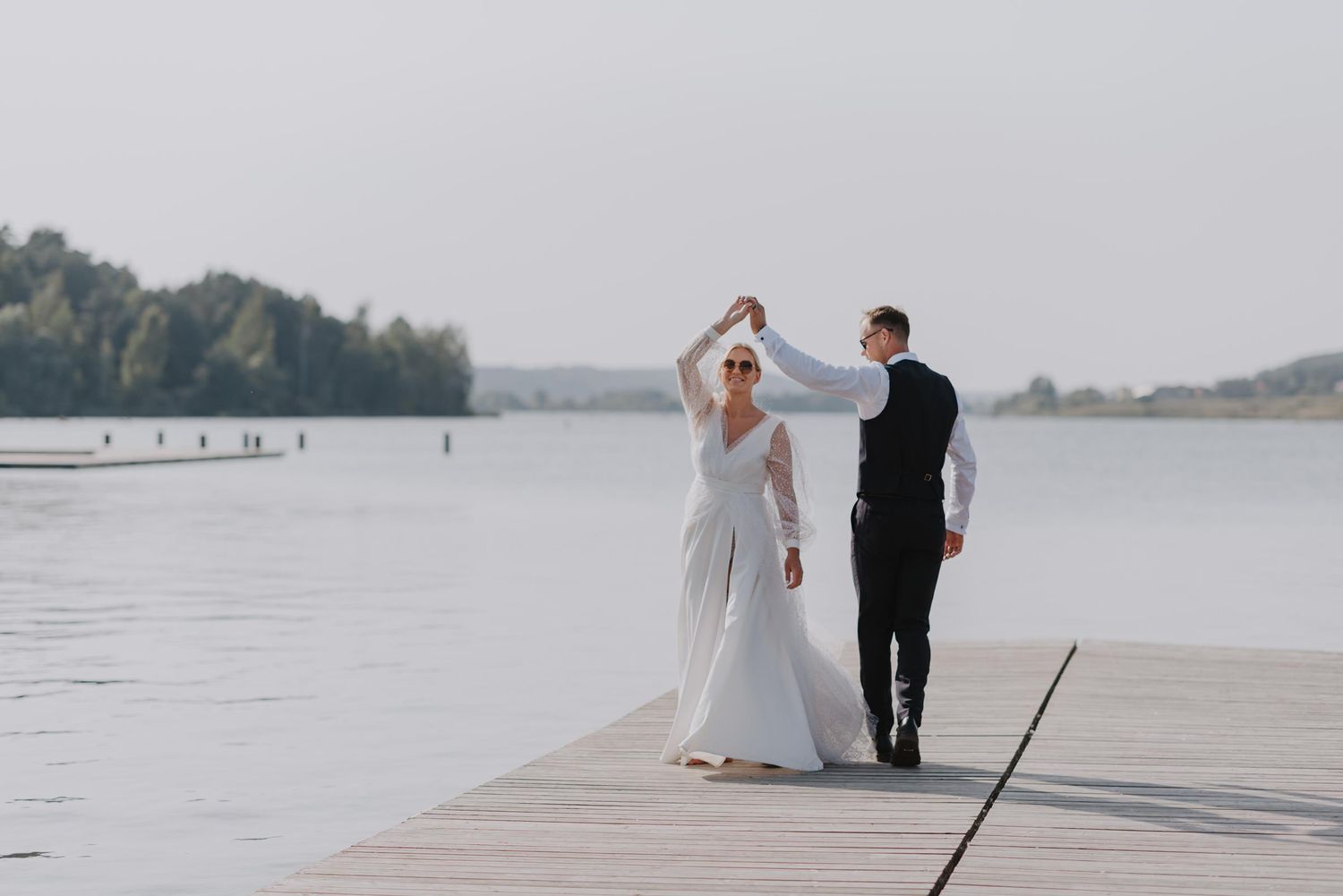 Couple walks along lakeside dock during wedding photoshoot.