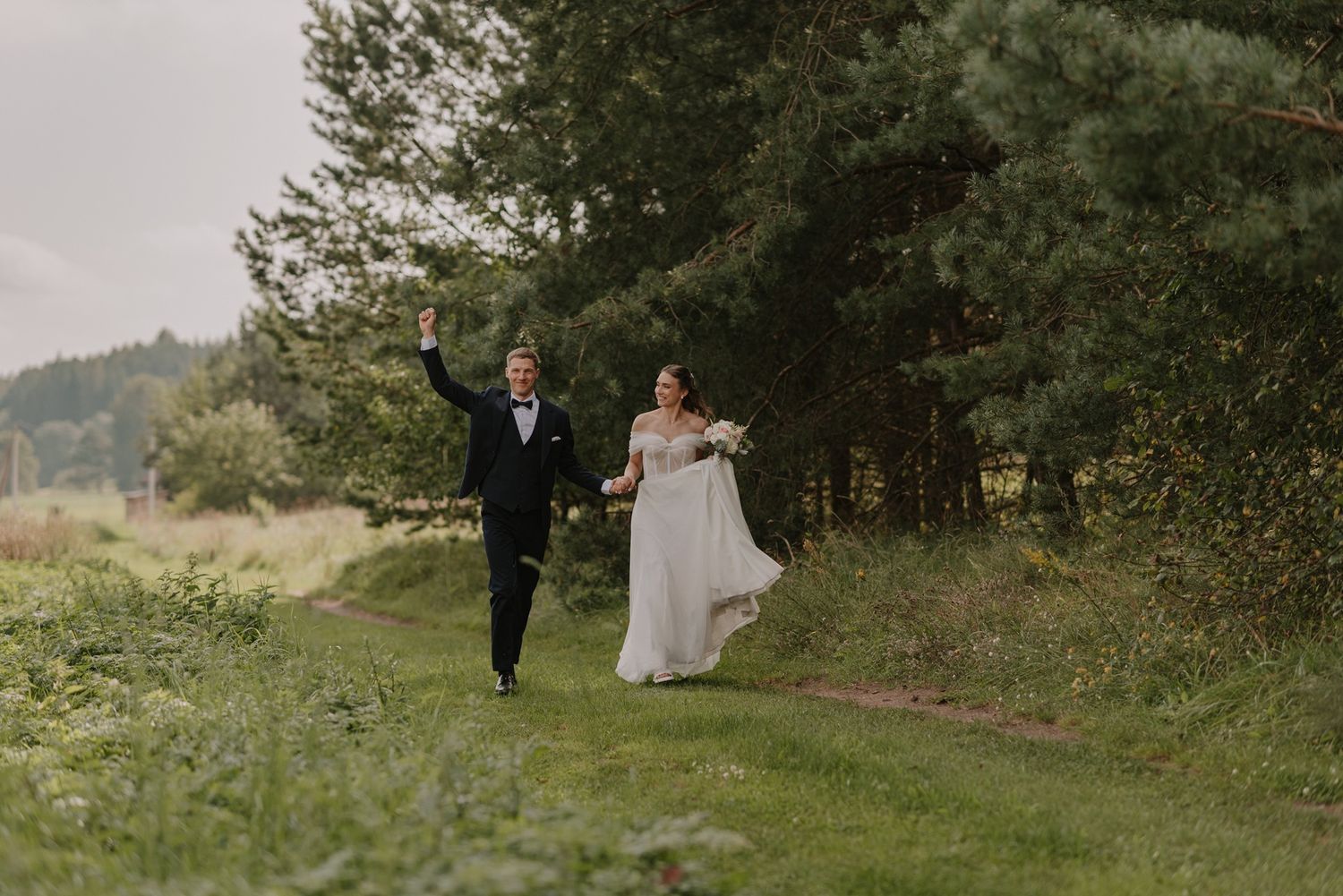 A couple in wedding attire walking together on a grassy path through a wooded area.