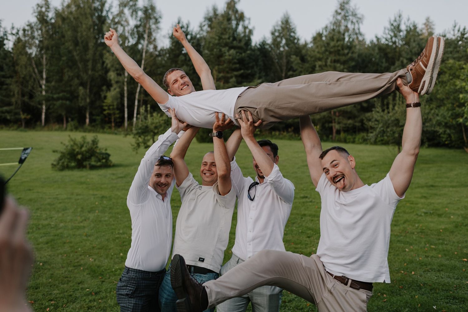 Wedding party celebrating joyfully by lifting someone in white clothing on a grassy lawn.