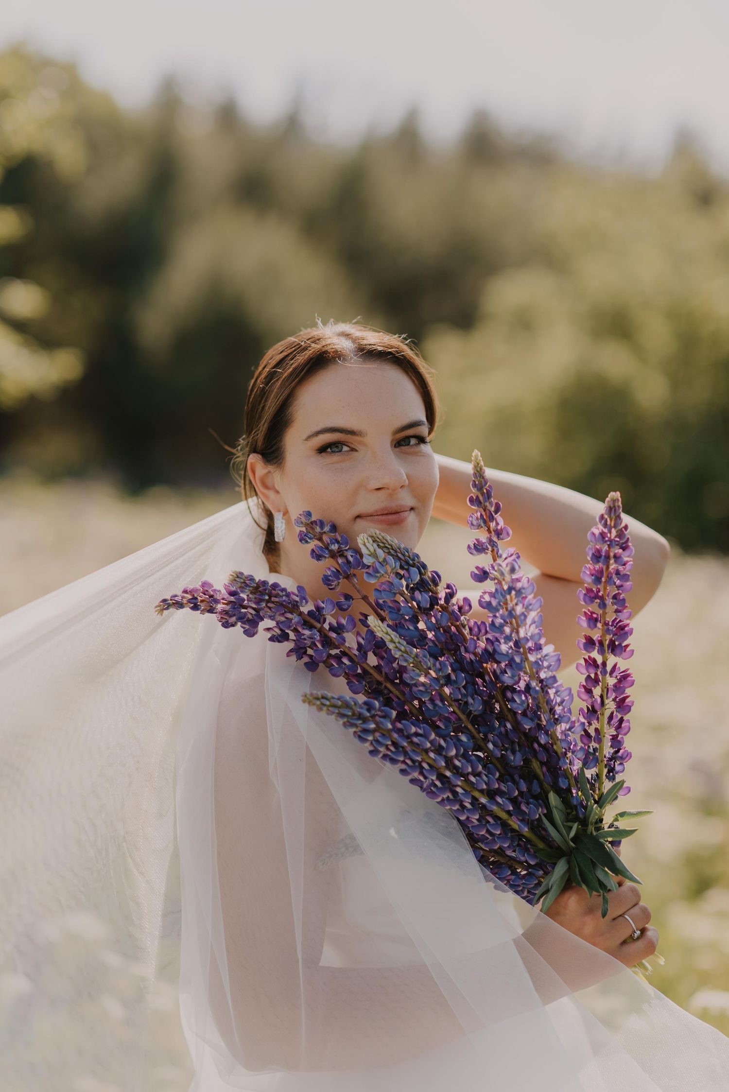 Artistic bridal portrait with purple lupine flowers and white veil in natural light.
