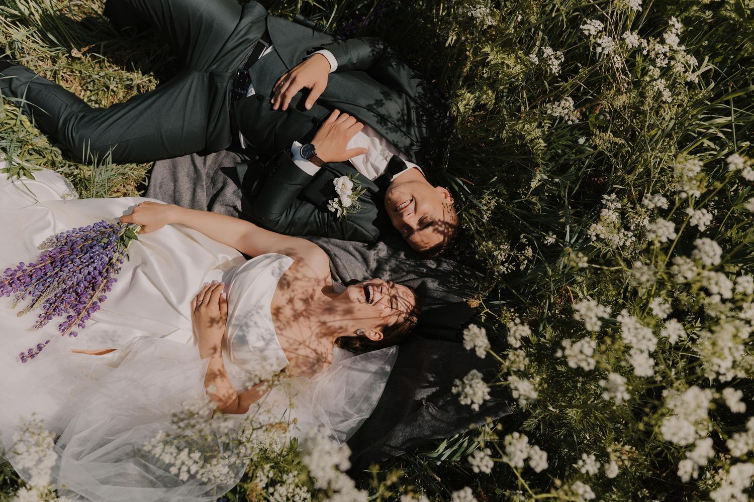 Couple lying in field surrounded by white flowers during wedding photo session.