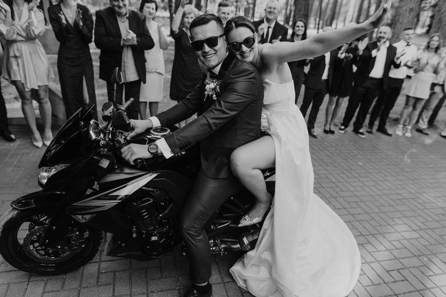 Wedding guests celebrate with the couple beside a motorcycle outdoors.