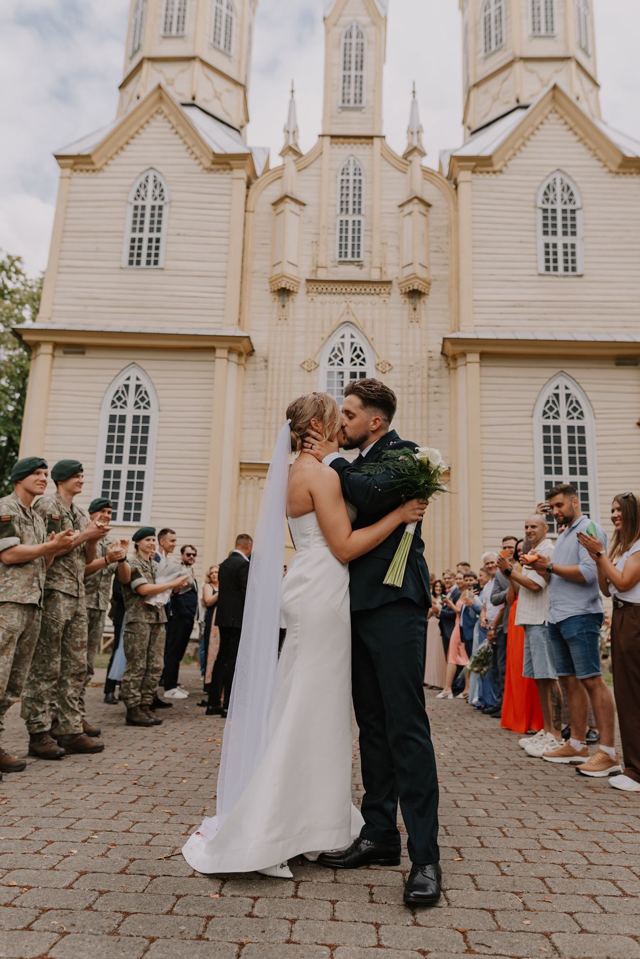 Couple celebrates their wedding in front of a beautiful white church with guests and military members present.