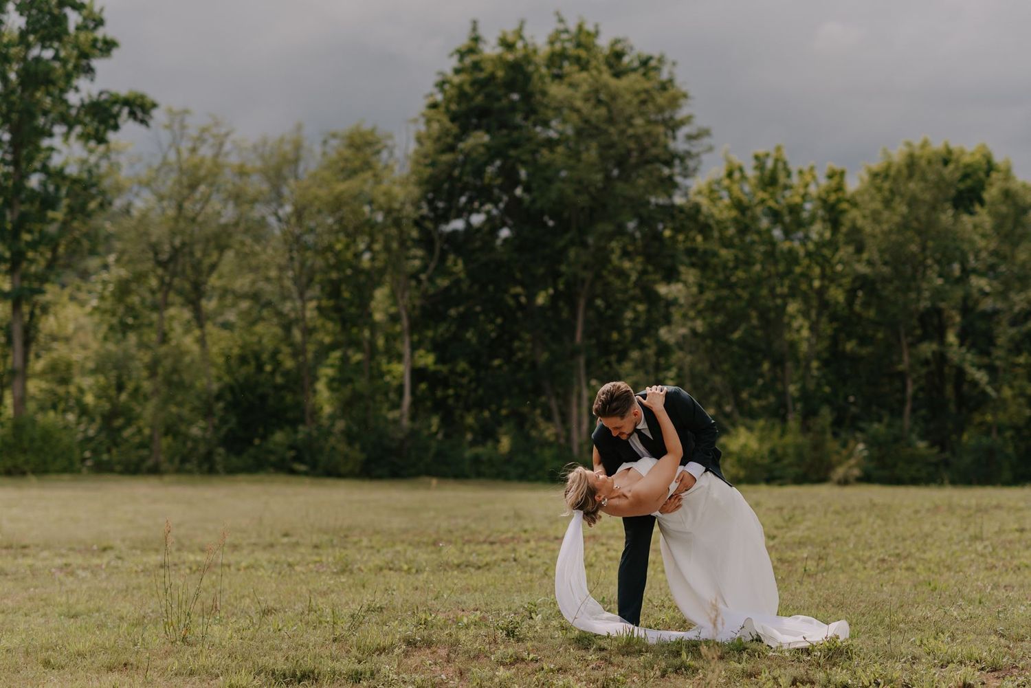 A wedding couple embraces in a scenic field with trees and moody skies in the background.