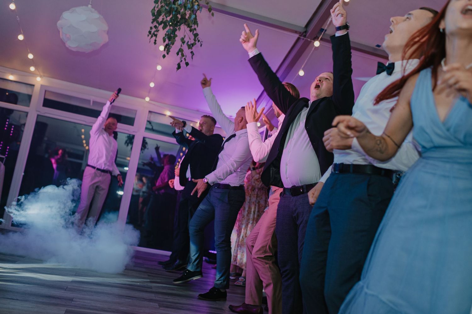 Wedding guests dance enthusiastically under purple lighting with atmospheric fog effects.