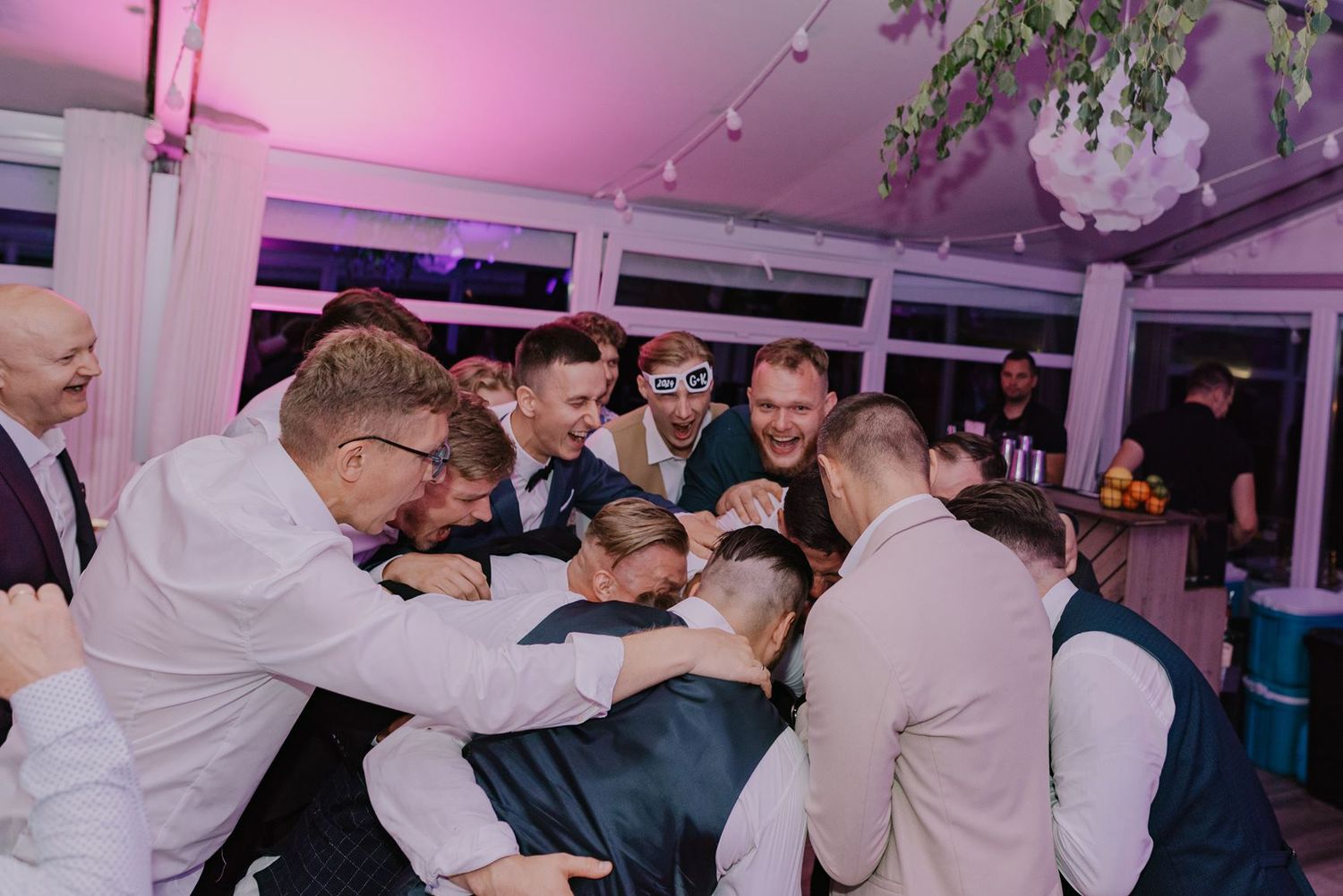 Wedding party dancing with hands raised in celebration under magenta lighting at reception.