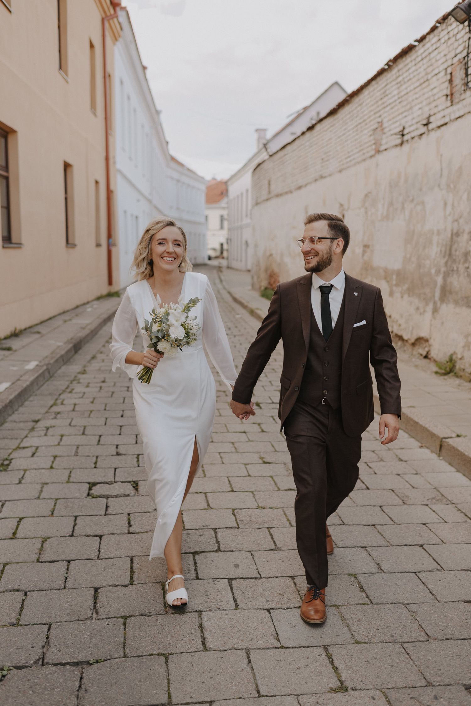Wedding couple strolling down a cobblestone path between cream colored buildings.