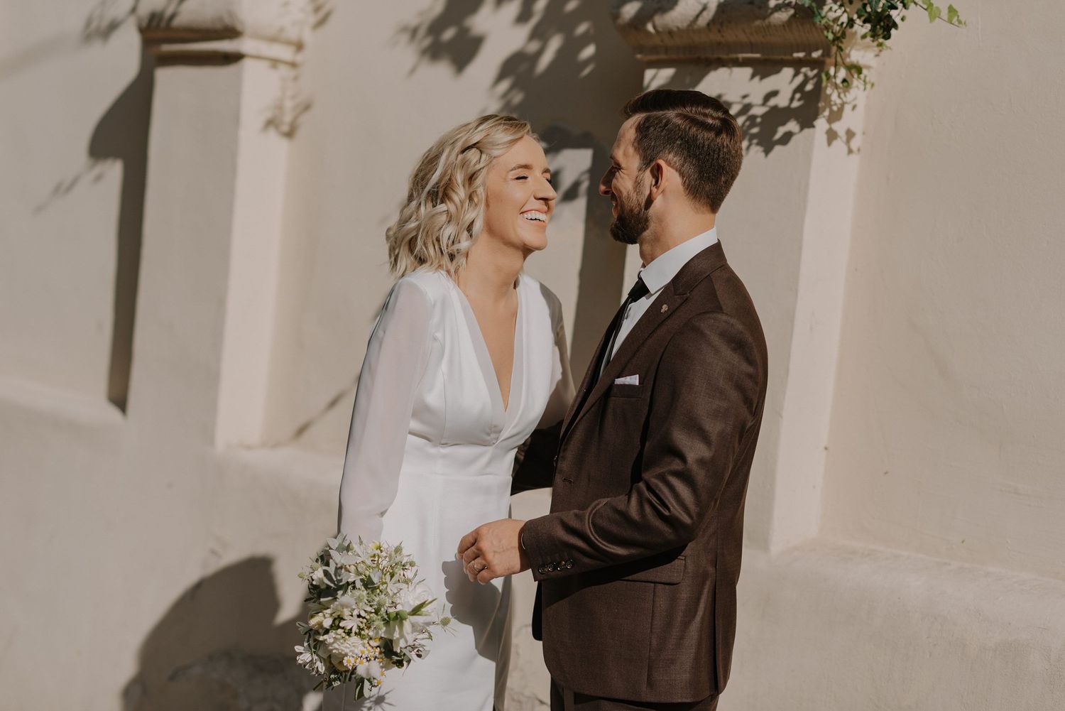 Intimate wedding portraits captured against a light stone wall in natural sunlight.