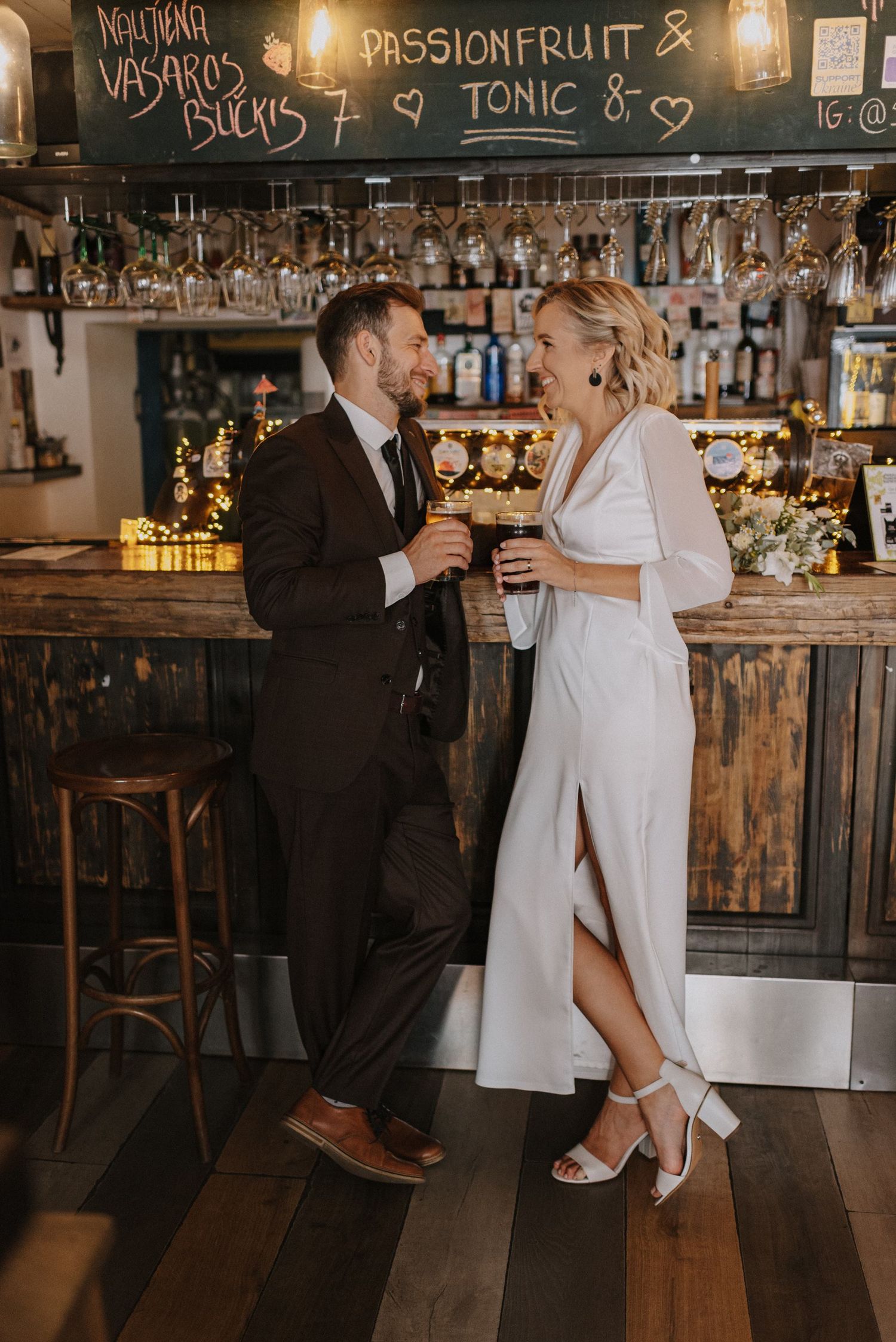 Wedding couple enjoying drinks at a rustic bar with wooden decor and chalkboard menu.
