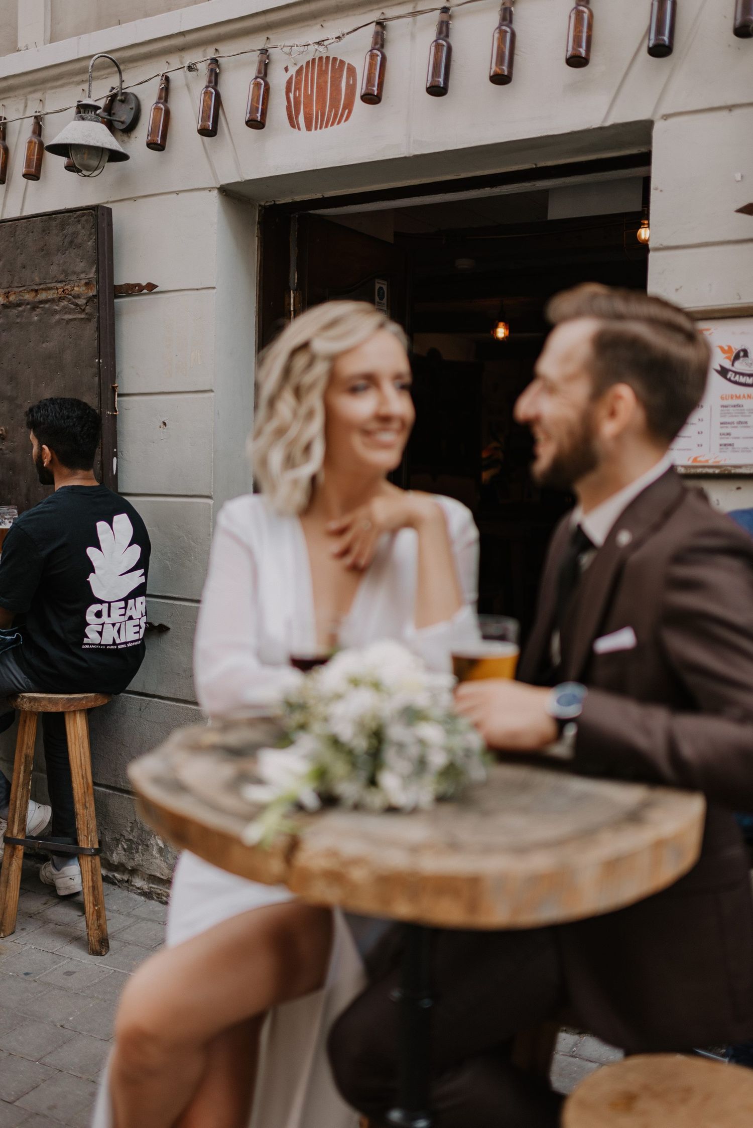 Newlyweds sharing moments at an outdoor cafe with hanging bottle decorations.