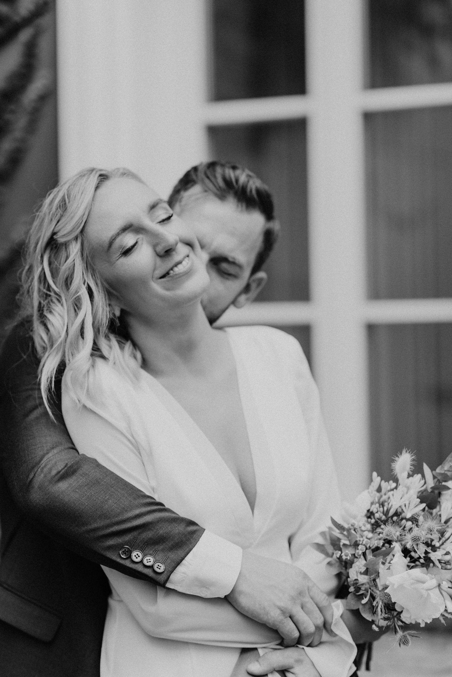 Black and white intimate wedding portrait capturing a romantic moment between newlyweds with a bridal bouquet.