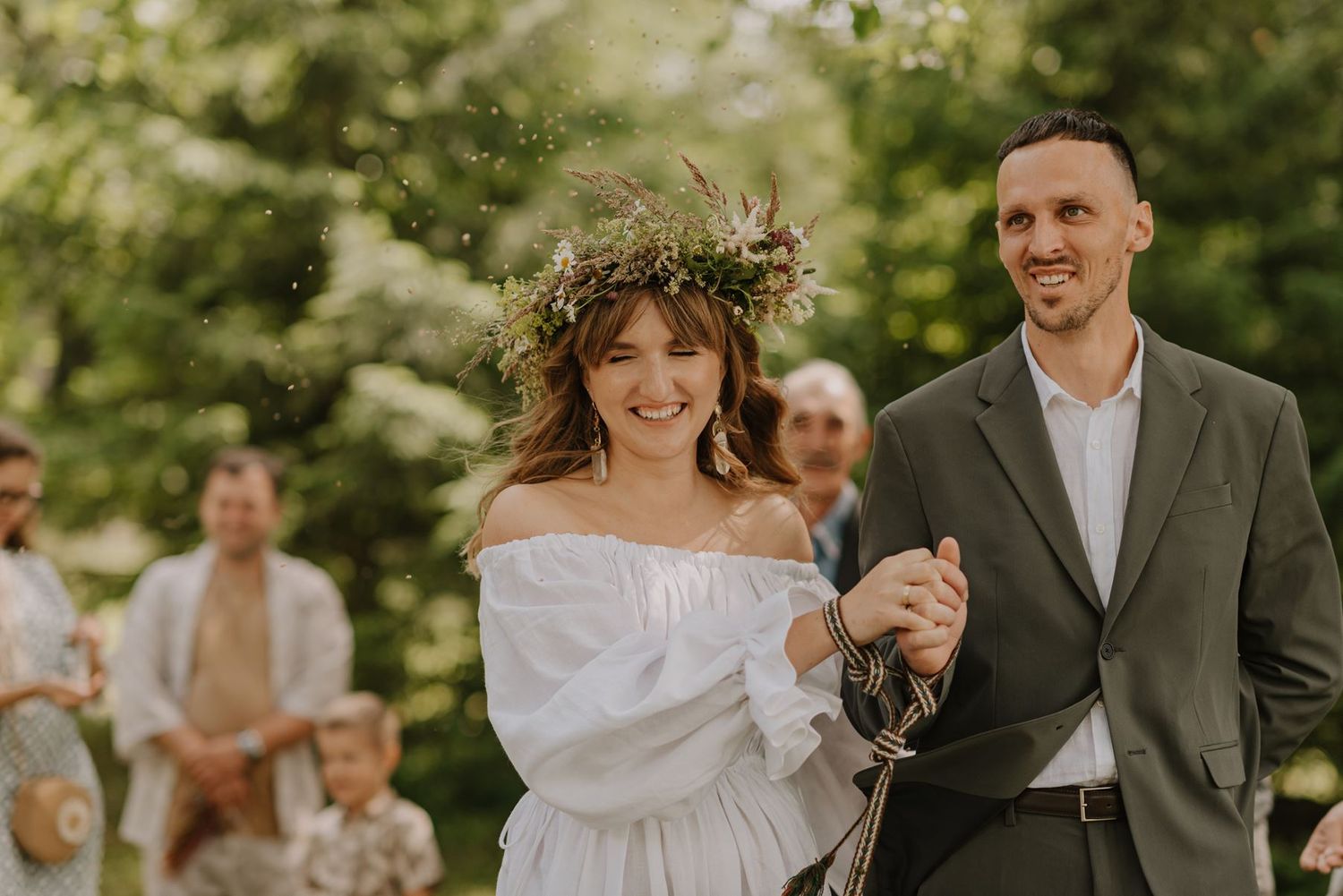 Joyful newlyweds walking outdoors during wedding celebration with flower crown and white dress.