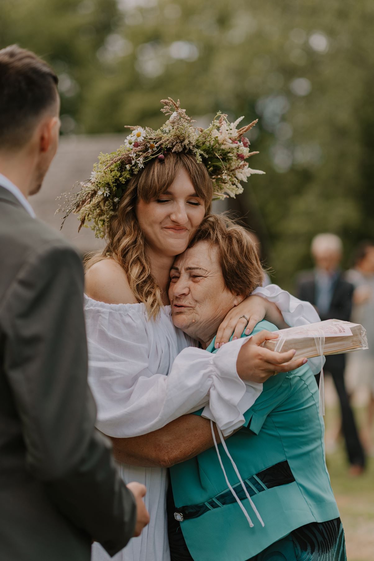 Two people embracing while wearing a floral crown and white dress in an outdoor setting.