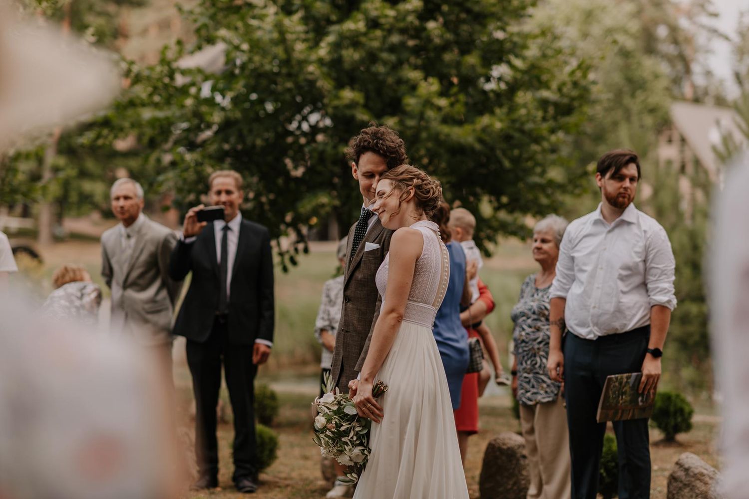 Wedding ceremony guests gathered outdoors under trees during a summer celebration.