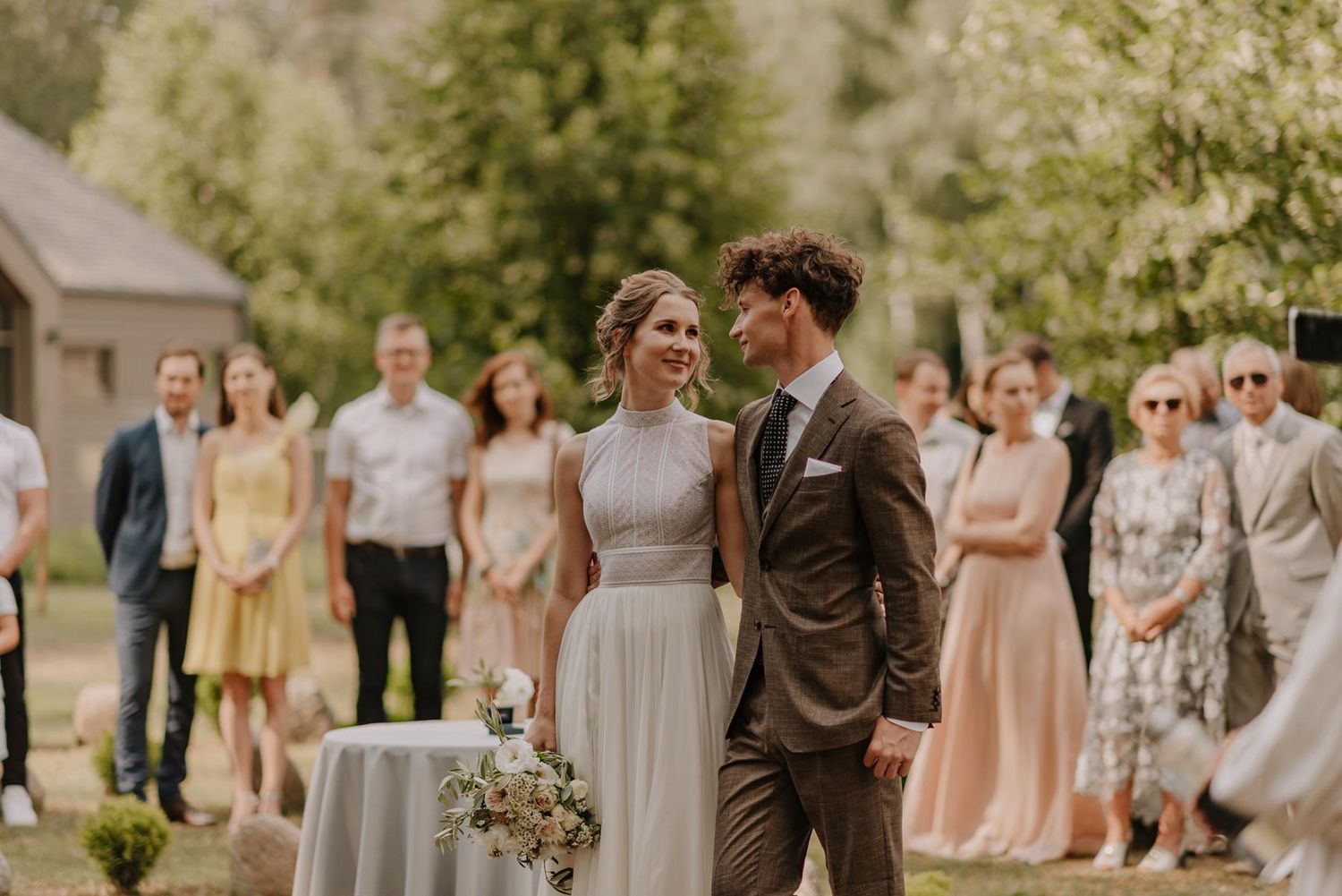 A couple stands together during their outdoor wedding ceremony surrounded by guests in pastel attire.