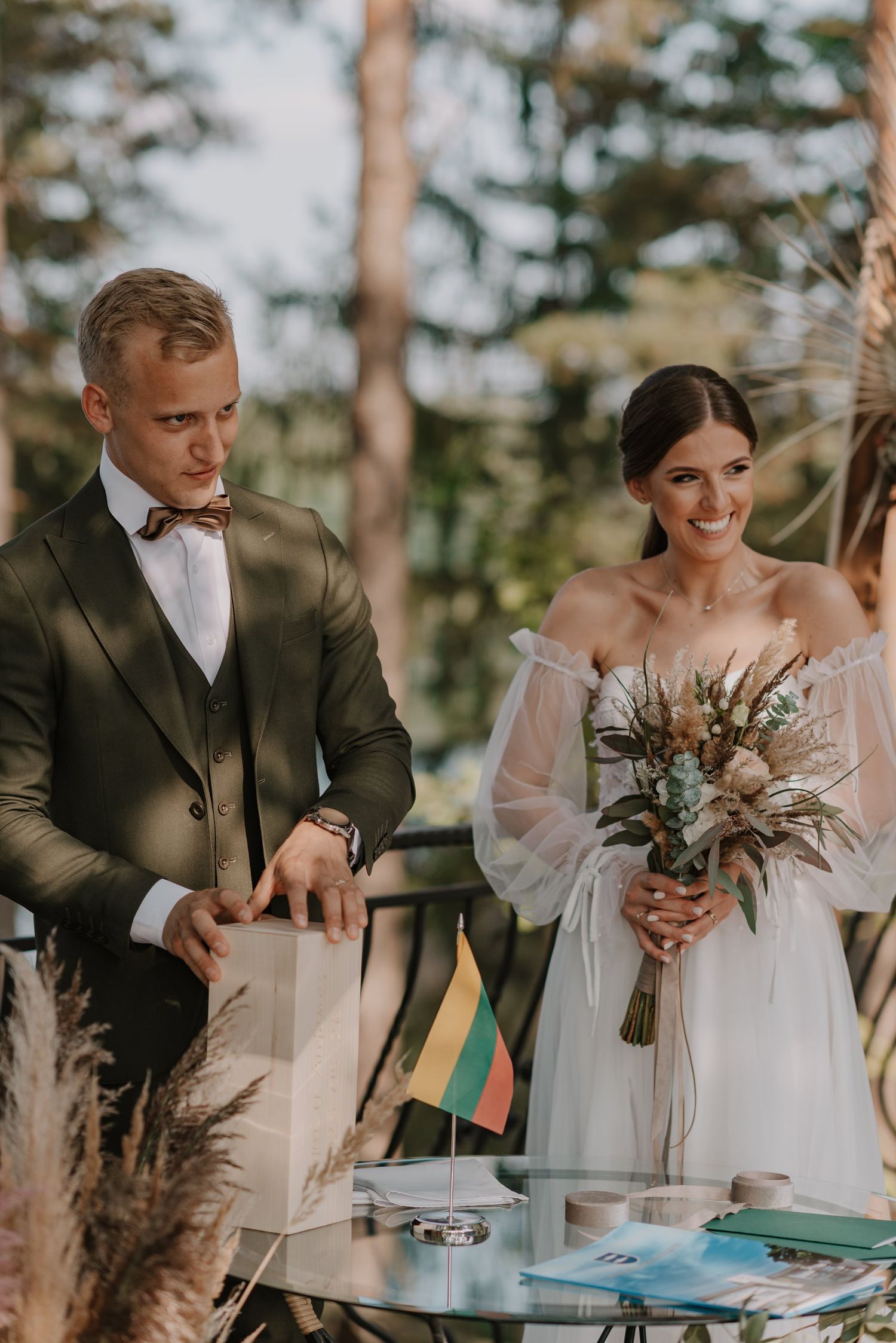 Couple signs marriage documents at rustic outdoor table with dried flower bouquet.