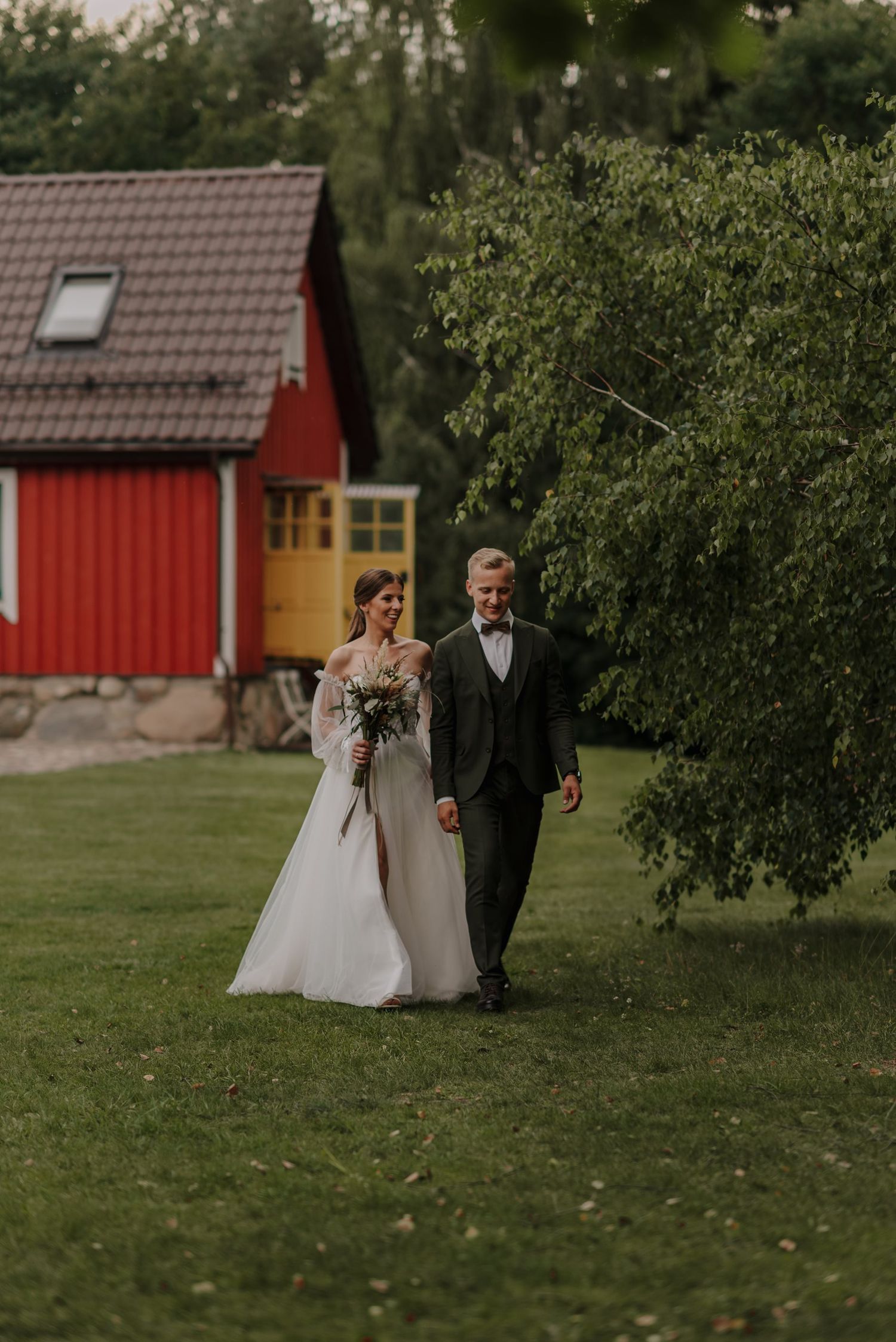 A couple walks together on their wedding day with a charming red house in the background.