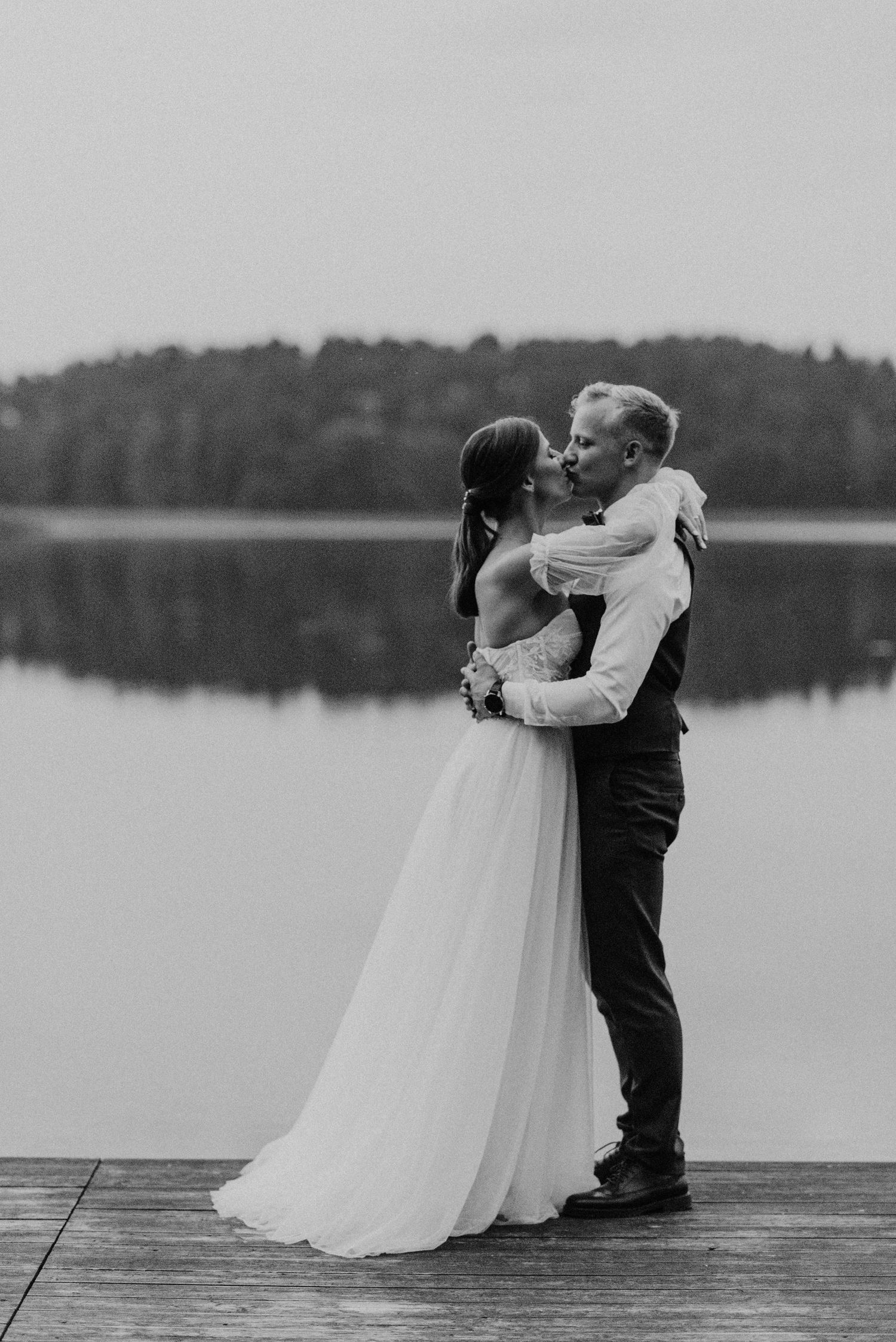 A romantic black and white wedding photo sequence of a couple embracing on a dock overlooking a serene lake.
