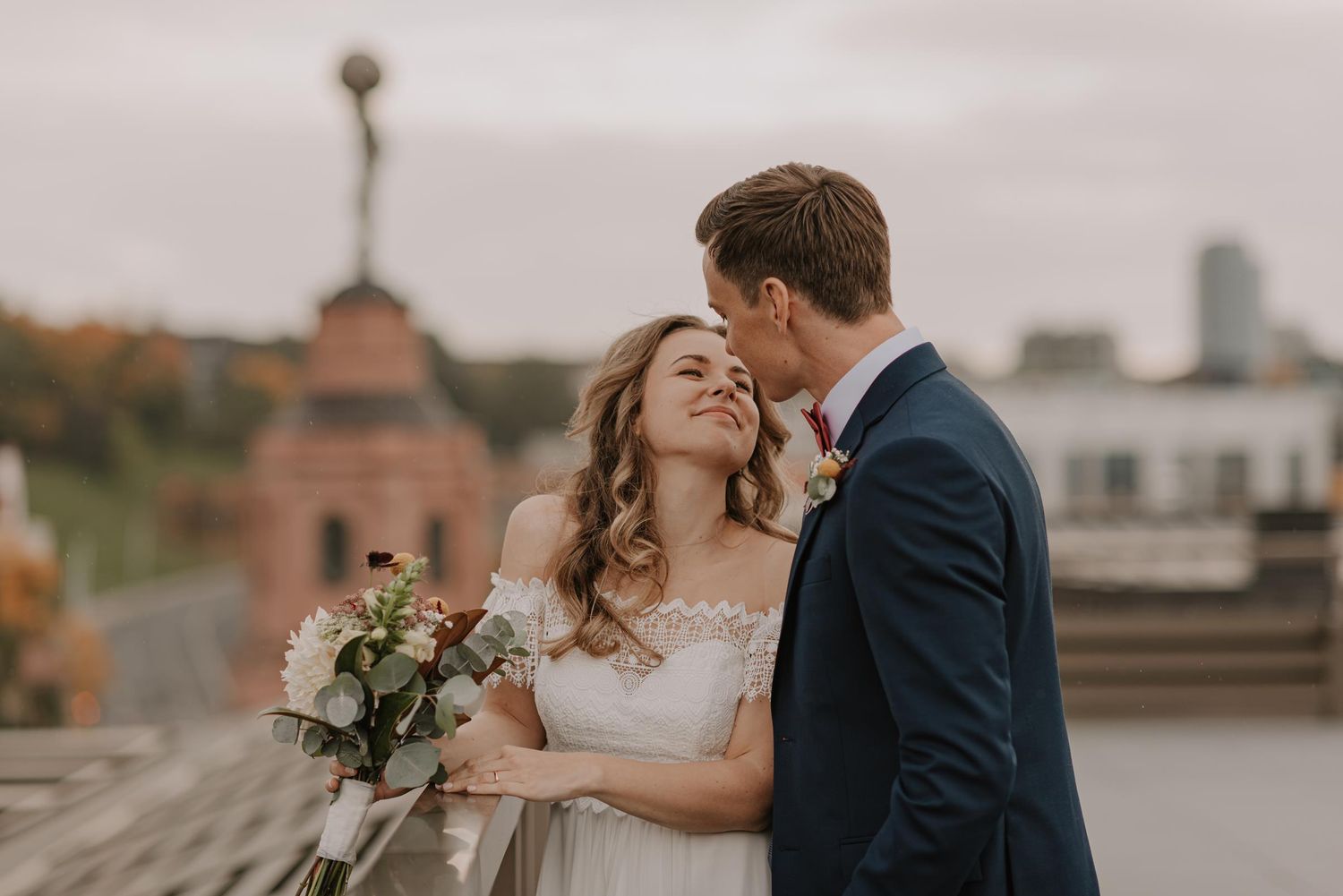 Newlyweds embrace during sunset on rooftop with city buildings in background.