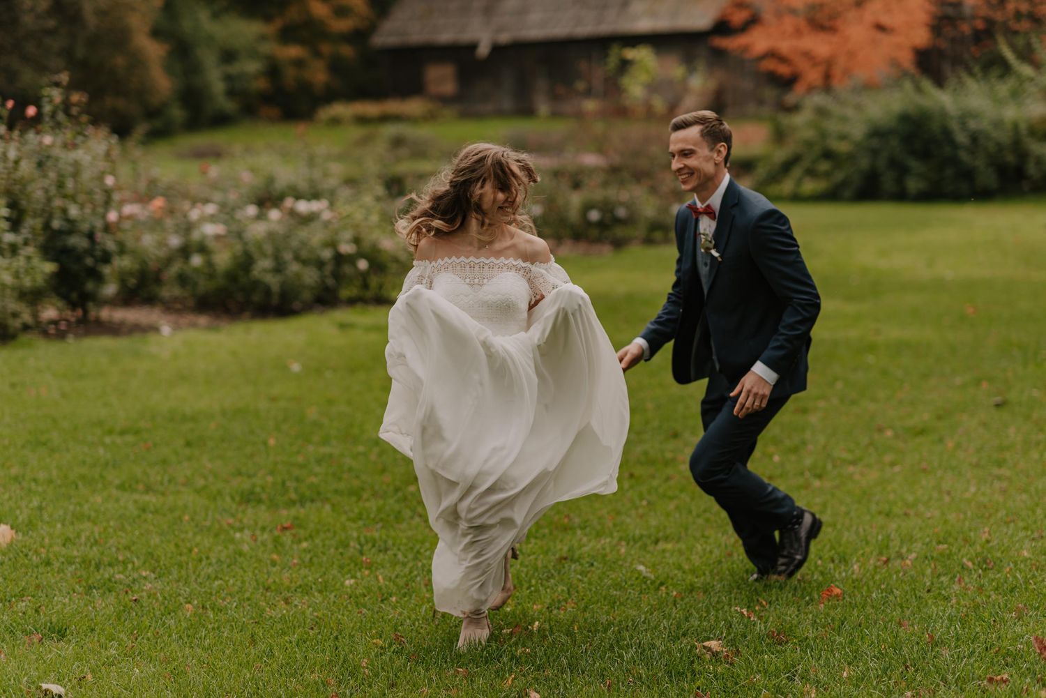 Newlyweds playfully run and dance across a lush green lawn during their autumn wedding celebration.