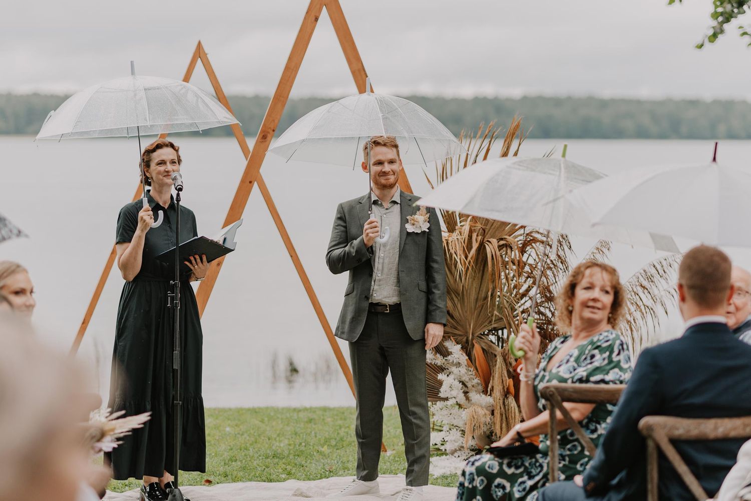 Wedding guests hold white umbrellas during an outdoor ceremony on a cloudy day.