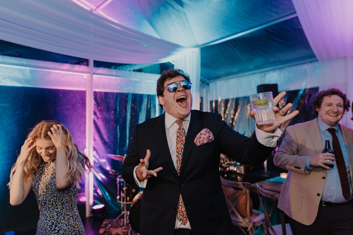Party guests celebrate with dramatic expressions and gestures under vibrant blue and purple lighting.