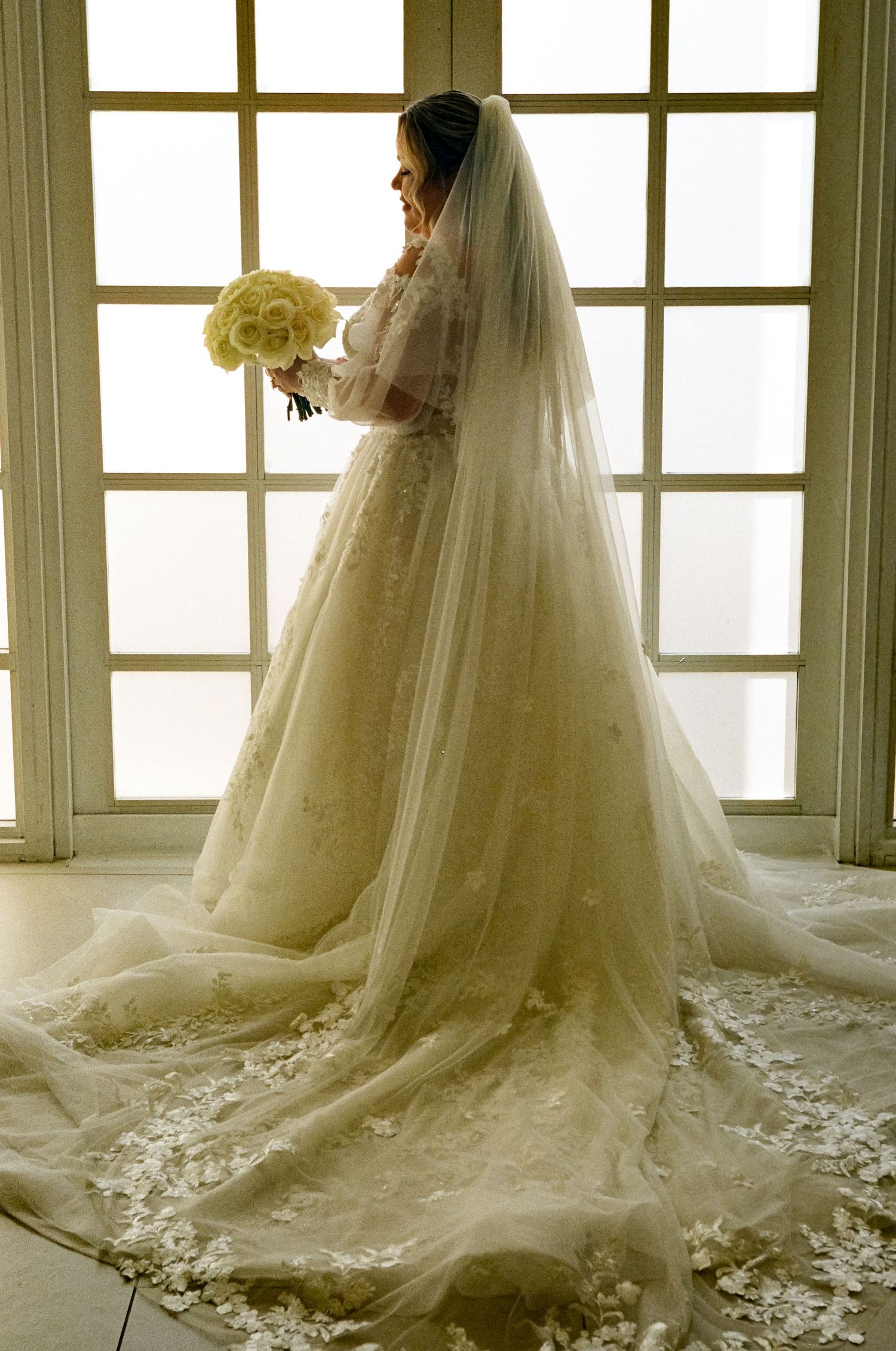 A bride in an elegant white wedding gown with a long flowing veil holds a cream-colored bouquet by a window.