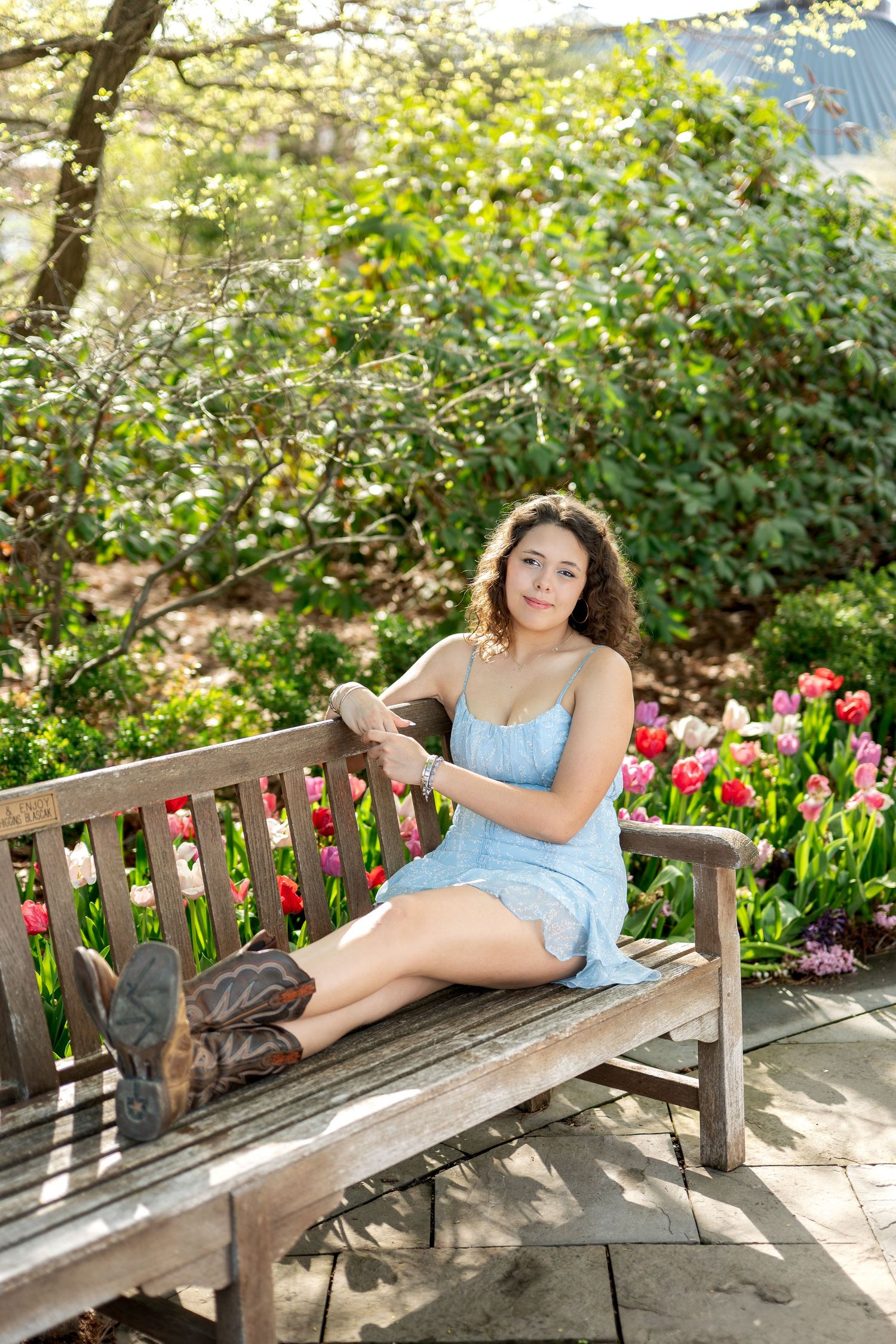 Young woman in blue dress and cowboy boots sitting on wooden bench surrounded by colorful tulips in a garden.