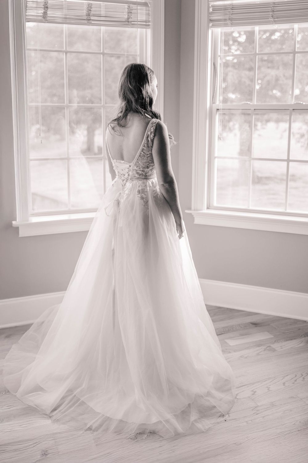 Back view of an elegant lace and tulle wedding gown in front of bright windows in a room with hardwood floors.