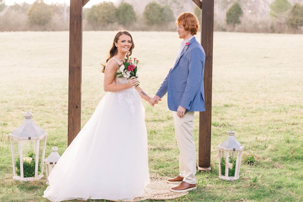 A rustic outdoor wedding ceremony under a wooden pergola decorated with white lanterns.