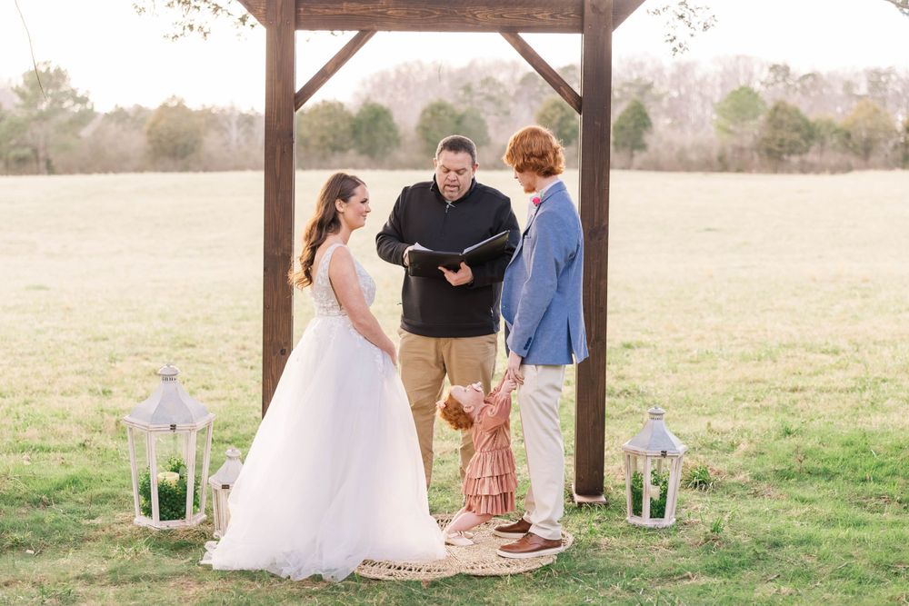 Wedding ceremony taking place under wooden arbor with lanterns in open field.