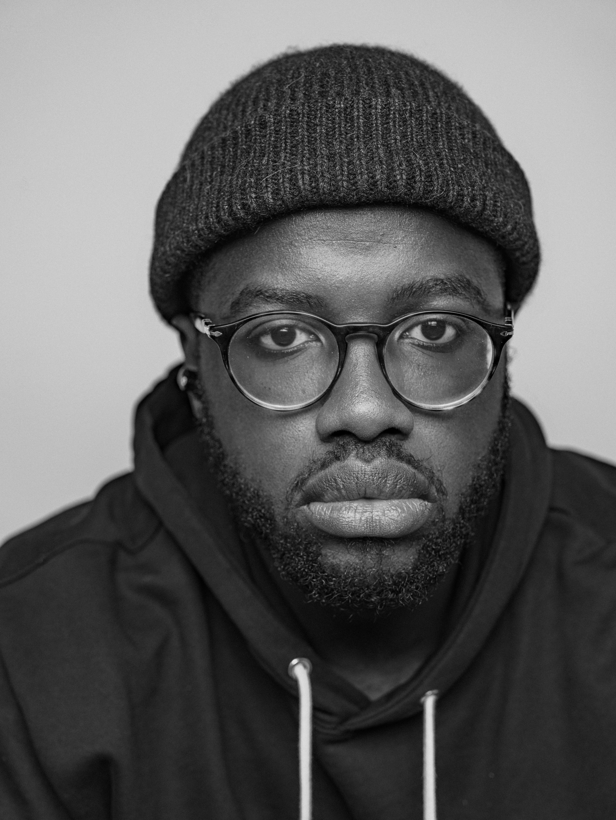 Portrait of a person wearing a knit beanie and glasses against a gray background.