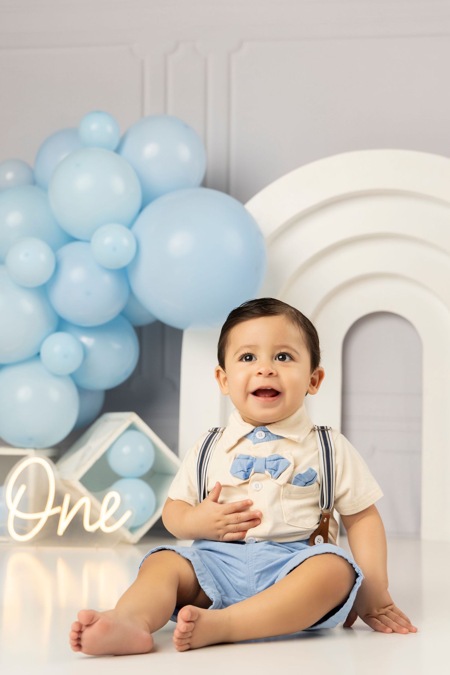 Baby boy in blue outfit sits smiling against white backdrop with blue balloon arch.