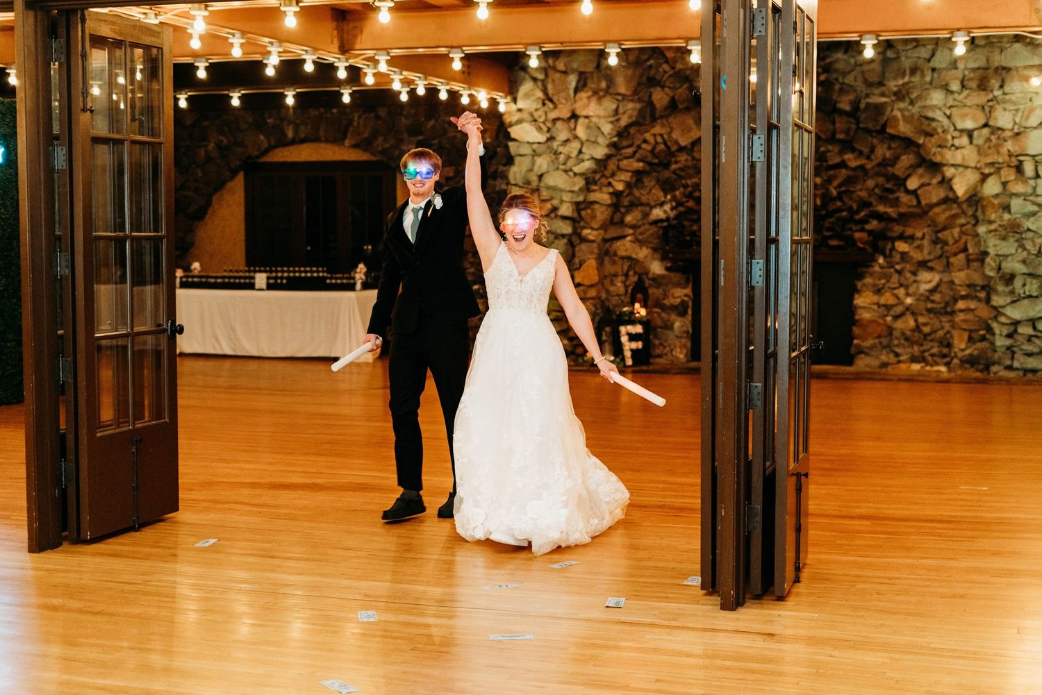 A joyful couple makes their wedding entrance through wooden doors into a rustic stone-walled venue with twinkling lights.