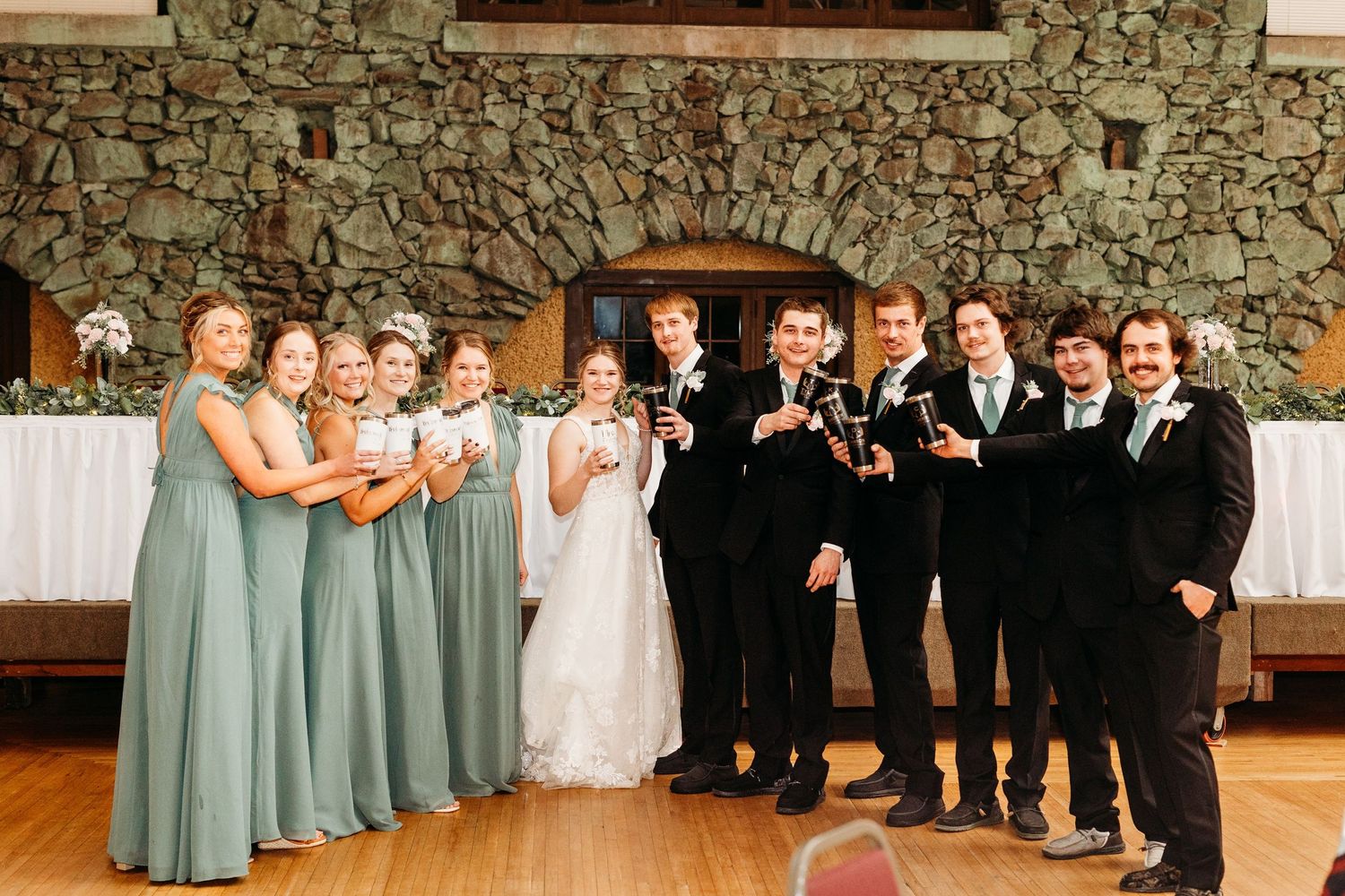 Wedding party in sage green dresses and black suits toasts with champagne glasses against a rustic stone wall.