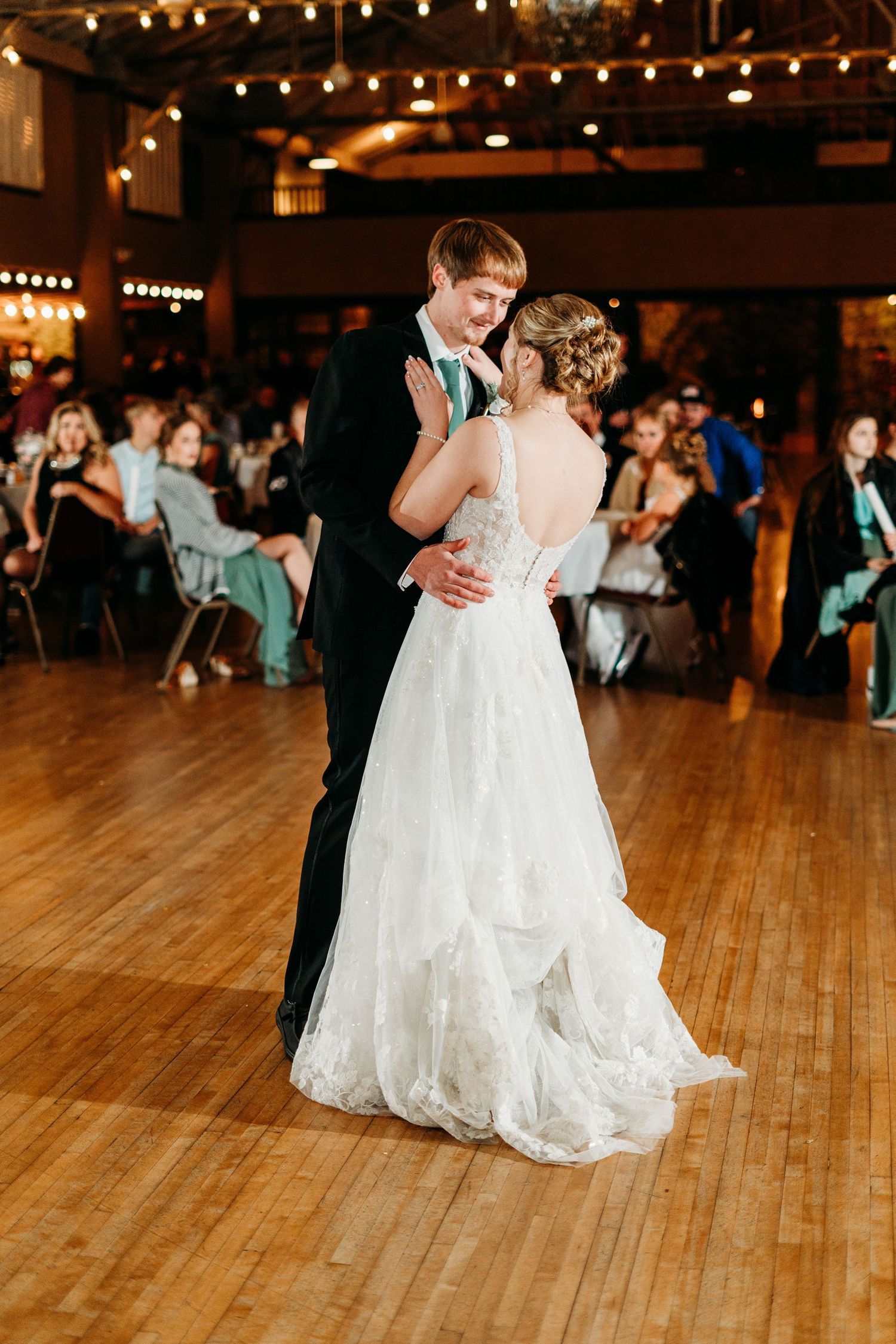 A couple share their first dance at their wedding reception on a wooden dance floor with string lights overhead.
