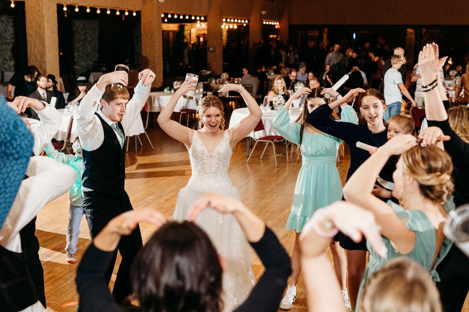 Wedding guests and the wedding party dance enthusiastically together on a wooden dance floor at the reception.