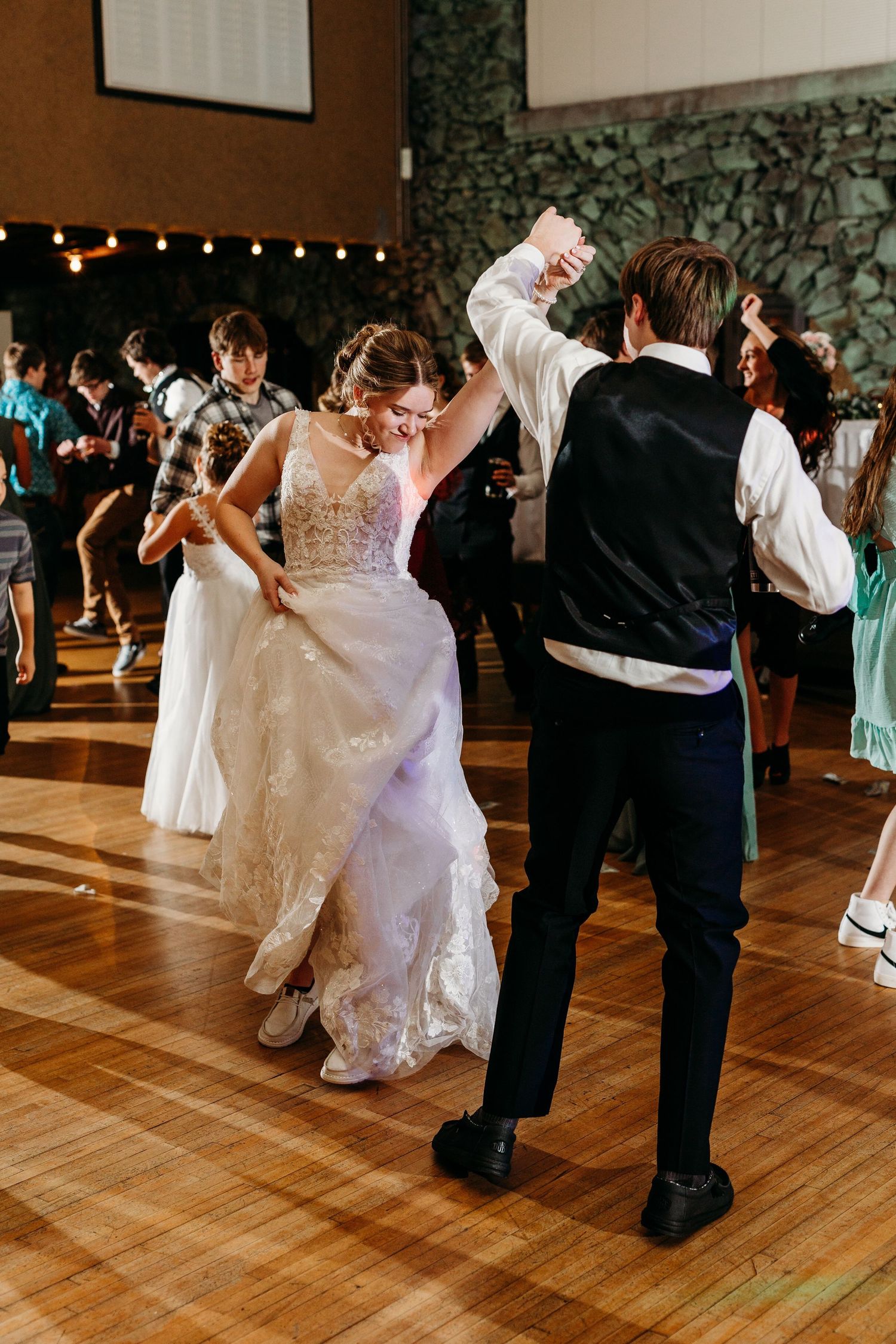 A bride in a flowing white lace dress dances with a partner on a hardwood floor during a wedding reception.