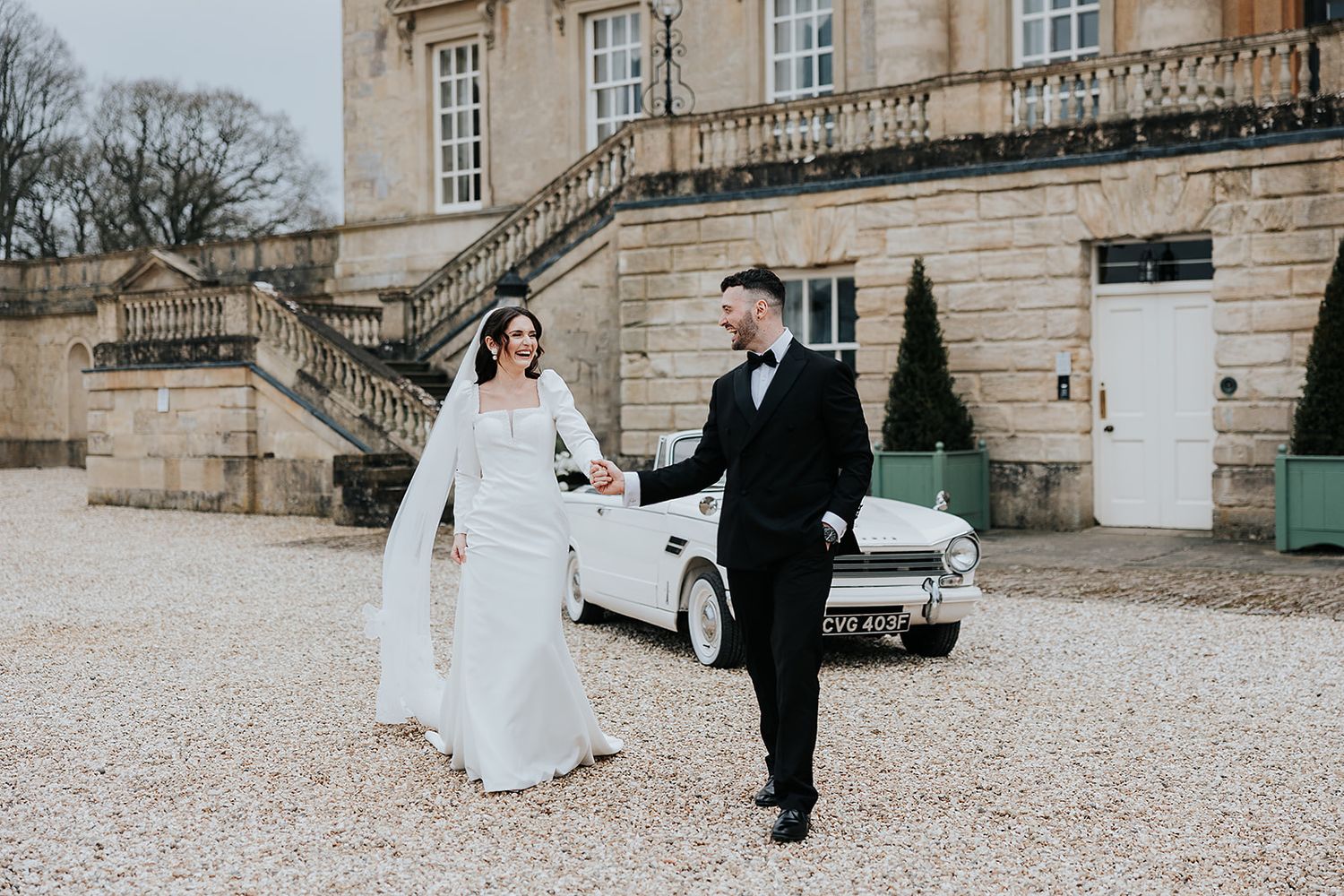 A couple in wedding attire walking hand in hand outside a historic mansion with a vintage white car in the background.