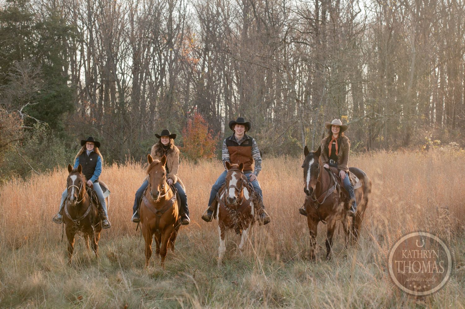 Four riders on horseback lined up in autumn field with bare trees behind them.