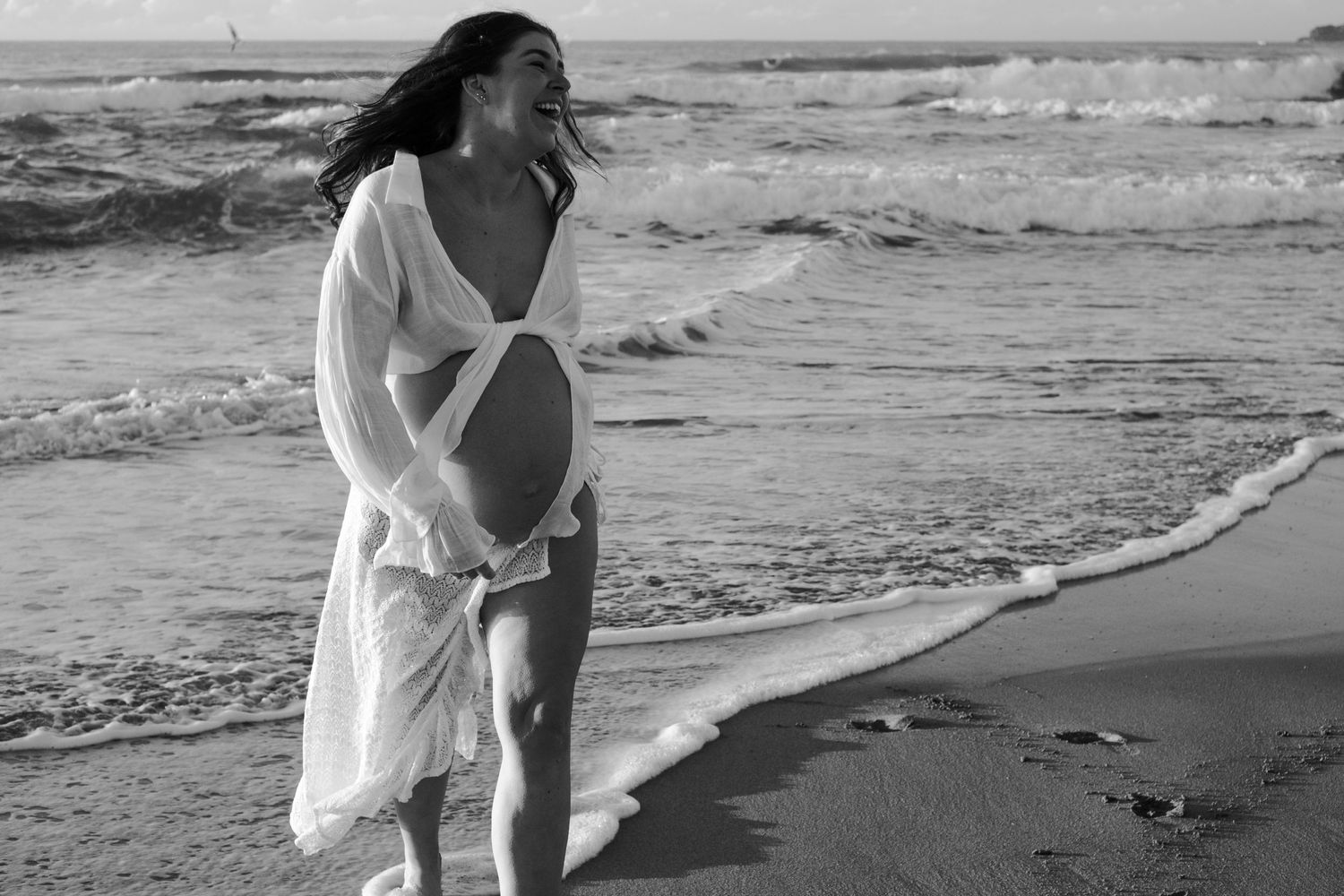 Pregnant woman in white dress walking along beach shore at sunset.