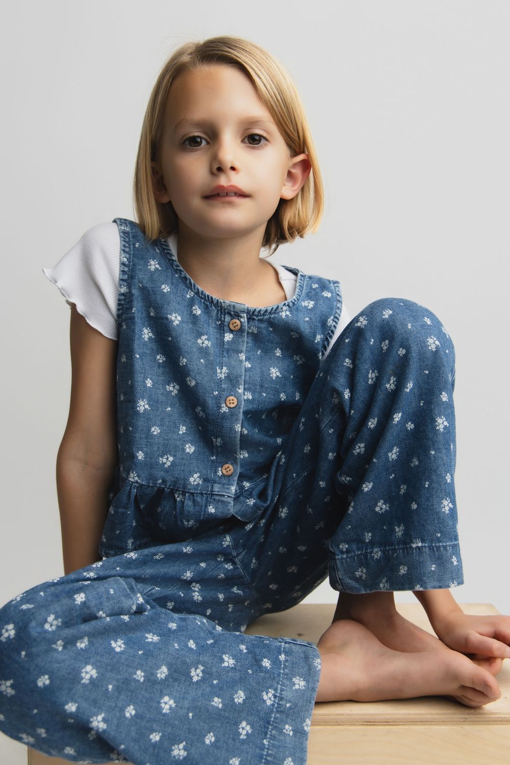 Child wearing a denim floral print jumpsuit with white sleeves sits casually on a wooden surface.