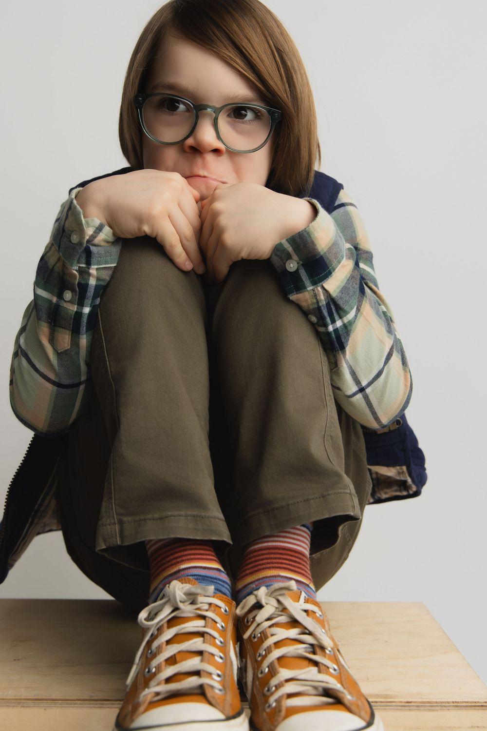 A young child with glasses sits hugging their knees, wearing a plaid shirt, khaki pants, striped socks, and yellow sneakers.