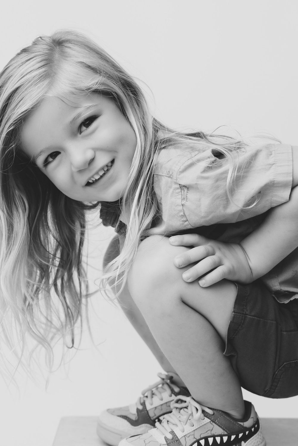 Black and white portrait of a smiling young blonde girl crouching playfully in casual clothes and sneakers.