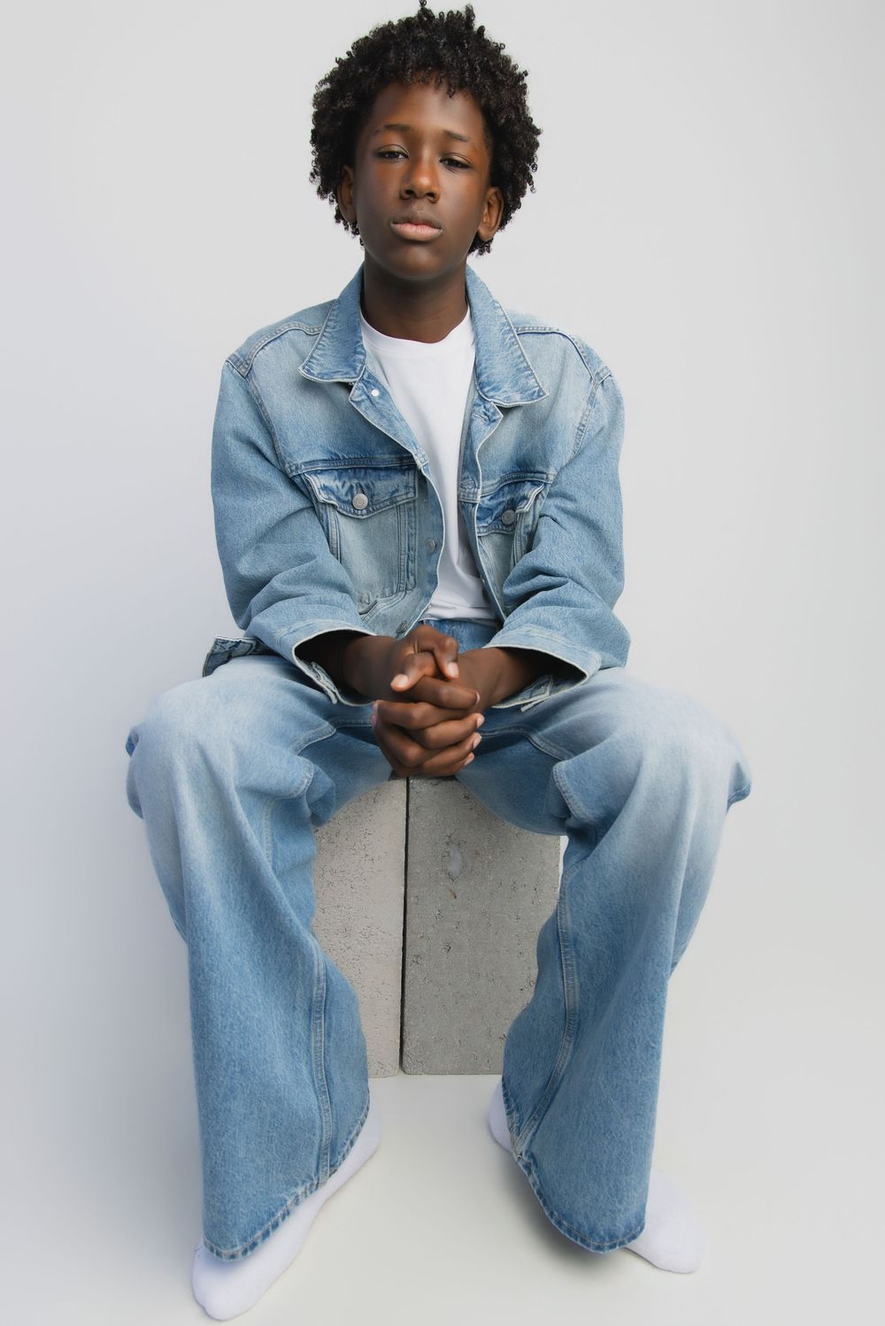 Young Black teen sitting on concrete block wearing full denim outfit with jacket and wide-leg jeans against white background.
